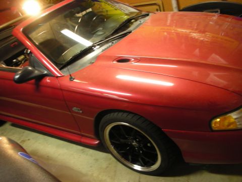 1998 Ford Crown Victoria - Angled front view of a red Mustang convertible with a hood scoop and black-and-chrome wheels, parked in a garage.