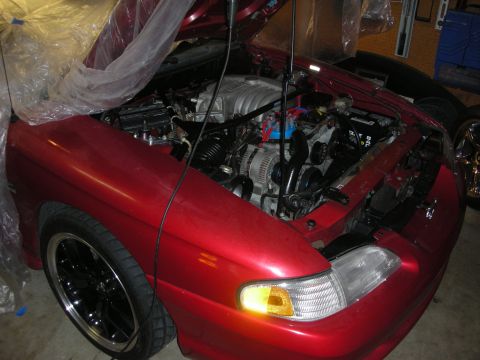 1998 Ford Crown Victoria - Open engine bay of a red car under work, showing the V6 engine, alternator and amber-lit front turn signal in a garage.
