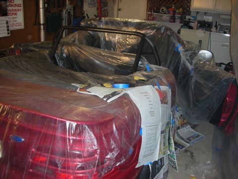 1998 Ford Crown Victoria - red convertible masked in plastic sheeting with an exposed roll bar and paperwork laid on the trunk in a garage.