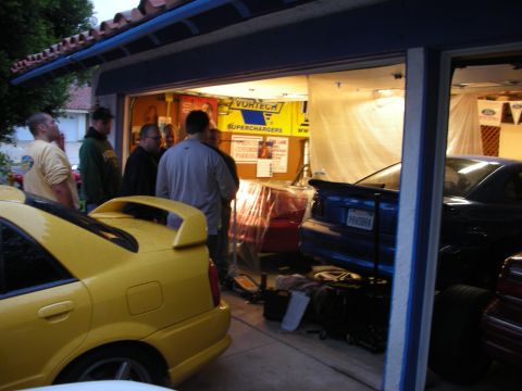 1998 Ford Crown Victoria - People gather outside an open garage at dusk near a yellow sedan, under a Vortech Superchargers banner.