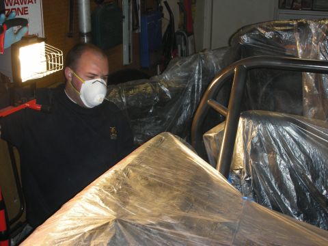 1998 Ford Crown Victoria - man wearing a white dust mask works beside a plastic-masked car and bare roll cage under a work light.