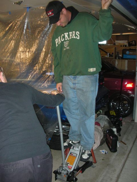 1998 Ford Crown Victoria - man in a green Packers sweatshirt and jeans stands atop a red floor jack in a garage while another person works nearby.