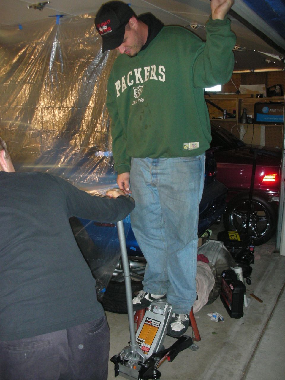 Man in green Packers sweatshirt standing on a floor jack in a garage