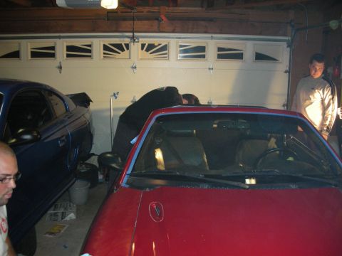 1998 Ford Crown Victoria - red convertible sits beside a darker car in a garage with a carriage-style door while two men look on at night.