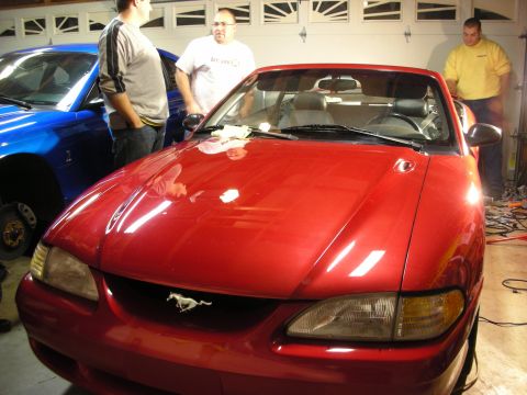 1998 Ford Crown Victoria - Front of a red Ford Mustang convertible bearing the running-pony grille emblem, with men standing around it in a garage.