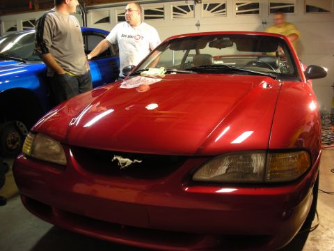1998 Ford Crown Victoria - front of a red Ford Mustang convertible with its pony grille emblem as men chat beside it in a lit garage.