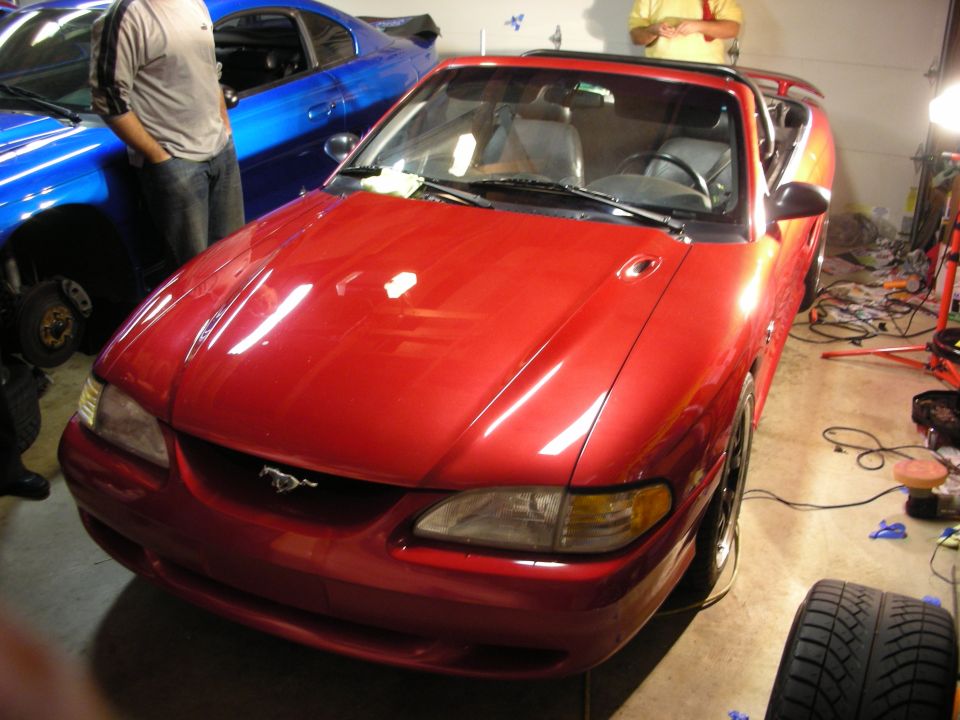 Red Mustang convertible front three-quarter view in garage with blue car nearby