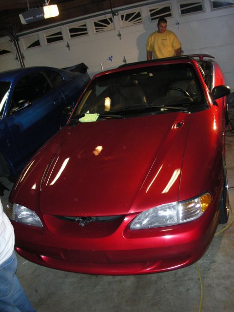 1998 Ford Crown Victoria - red Ford Mustang convertible shows its pony grille emblem in a garage as a man stands behind it near a blue car.