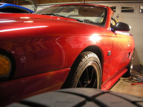 1998 Ford Crown Victoria - Side front view of a glossy red Mustang convertible showing its front fender, mirror and black mesh wheel in a garage.