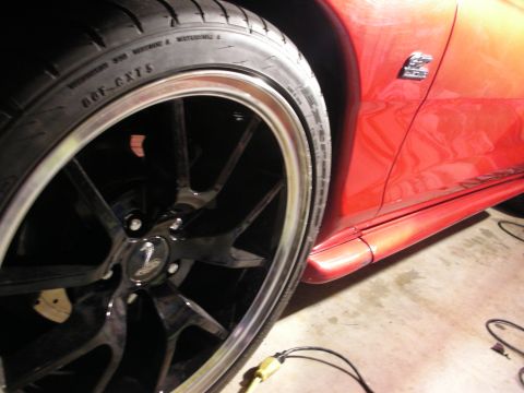 1998 Ford Crown Victoria - Detail of a glossy black multi-spoke wheel and low-profile tire on a metallic orange-red sports car parked in a dim garage.