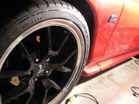 1998 Ford Crown Victoria - Close-up of an orange-red sports car's rear quarter showing a black multi-spoke wheel and performance tire under garage lighting.
