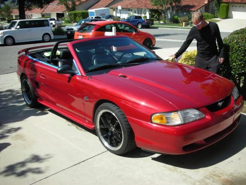 1998 Ford Crown Victoria - red Ford Mustang GT convertible with top down parked in a suburban driveway as a man stands at the passenger side.