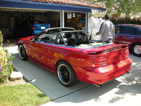 1998 Ford Crown Victoria - Rear three-quarter view of a red Ford Mustang convertible in a driveway near an open garage with a man working at the trunk.
