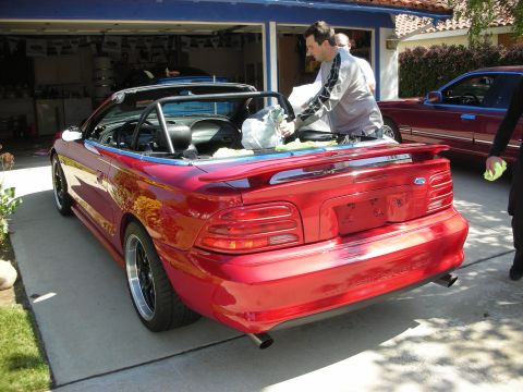1998 Ford Crown Victoria - Rear view of a red Ford Mustang convertible showing dual exhaust tips and trunk spoiler, with men working on it in a driveway.