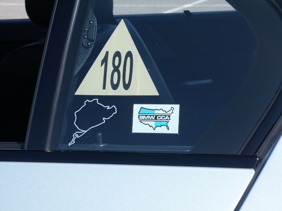 Car window with a yellow triangle sticker showing '180', a Nürburgring outline, and a BMW CCA map sticker