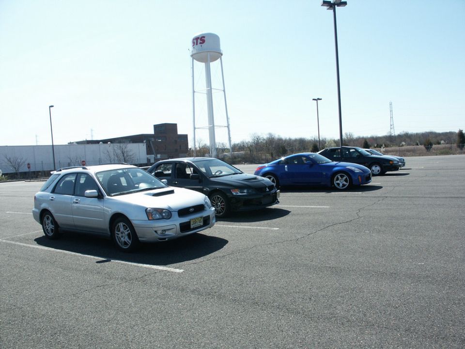 Parking lot scene with a silver Subaru Impreza wagon, black Mitsubishi Lancer Evolution, blue Nissan 350Z, and a dark sedan, with a water tower in the background