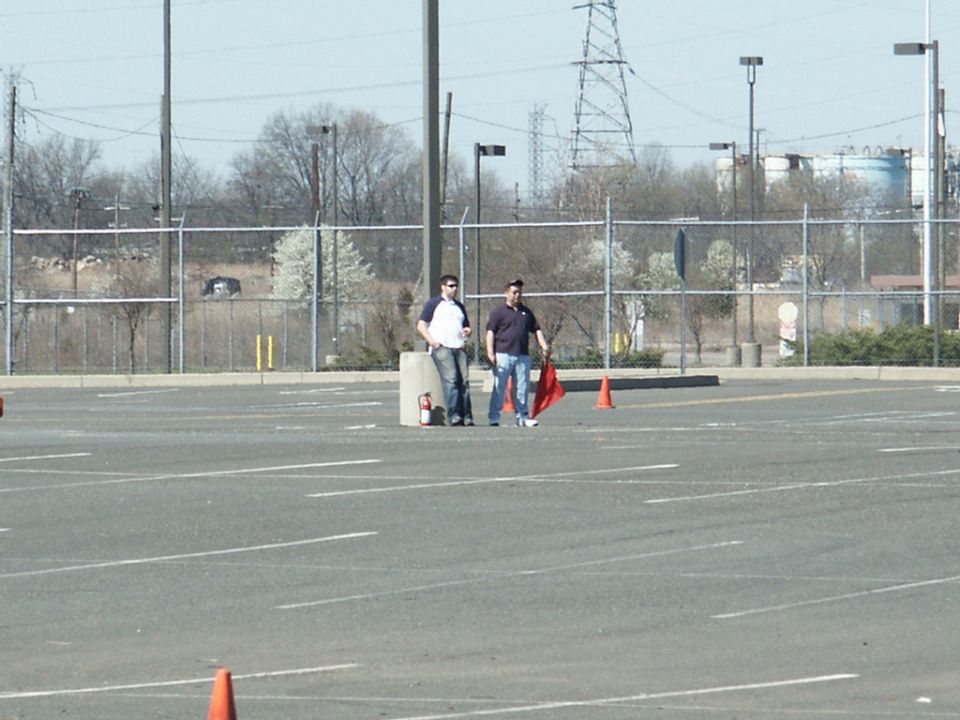 Two men stand in a paved lot with orange cones and a fire extinguisher