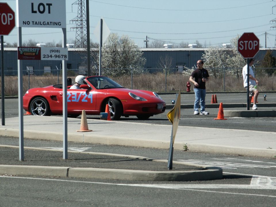 Red convertible sports car, possibly a Porsche Boxster, with "23/24" on the door, parked on a lot with cones