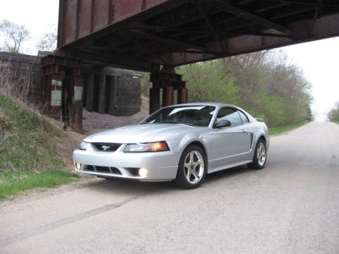 1998 Ford Taurus - A silver Ford Mustang coupe is parked on a gravel road under a large railway bridge.
