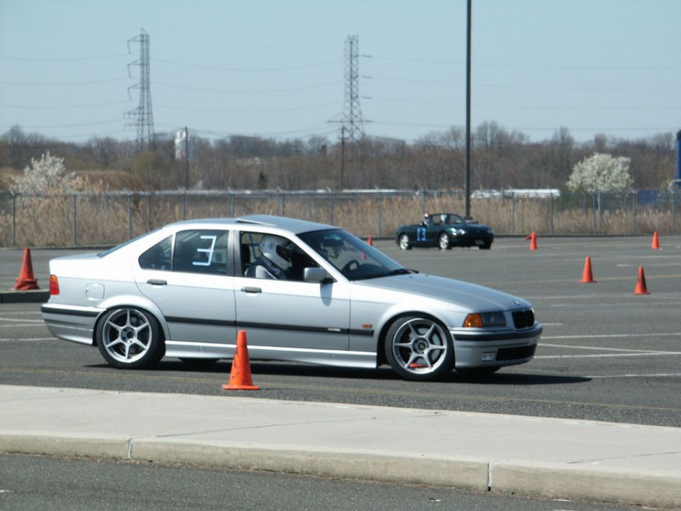 Silver BMW sedan with racing number 3 on door, driving on a track with orange cones. Black convertible in background