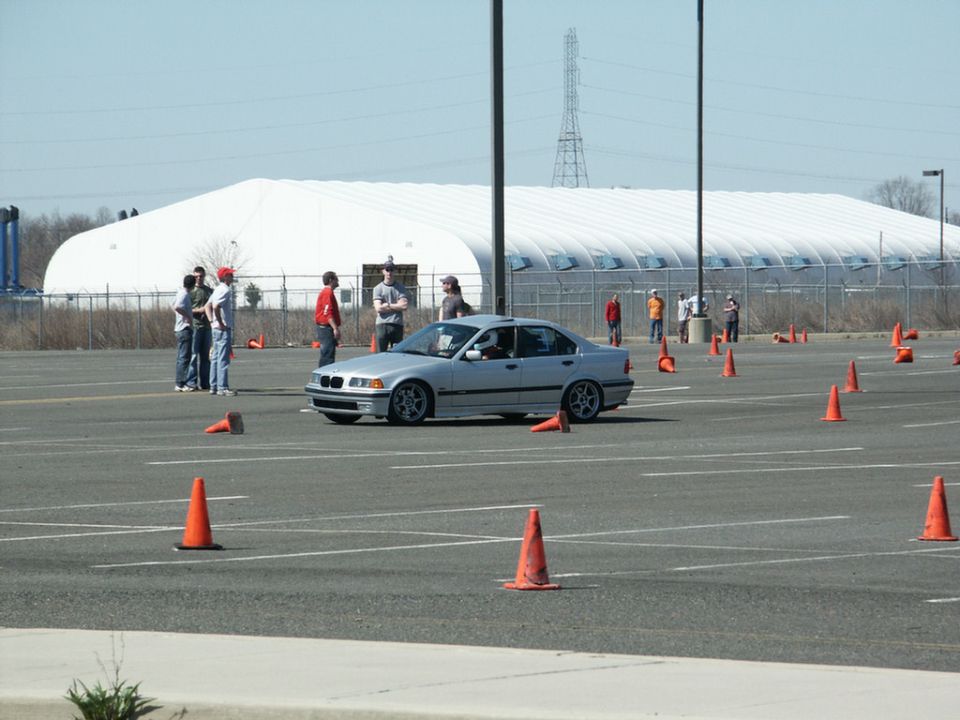 Silver BMW sedan navigating a course marked by orange cones, with people standing around in the background near a large white building
