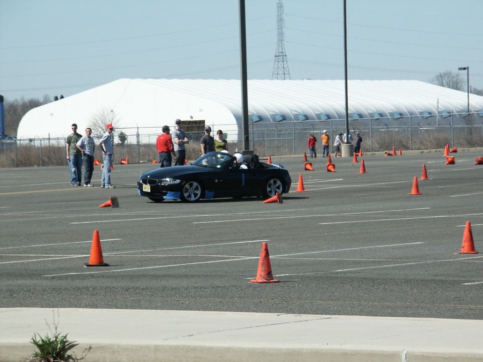 Black convertible sports car with blue trim on front bumper, parked on a lot with orange cones. Several people stand nearby