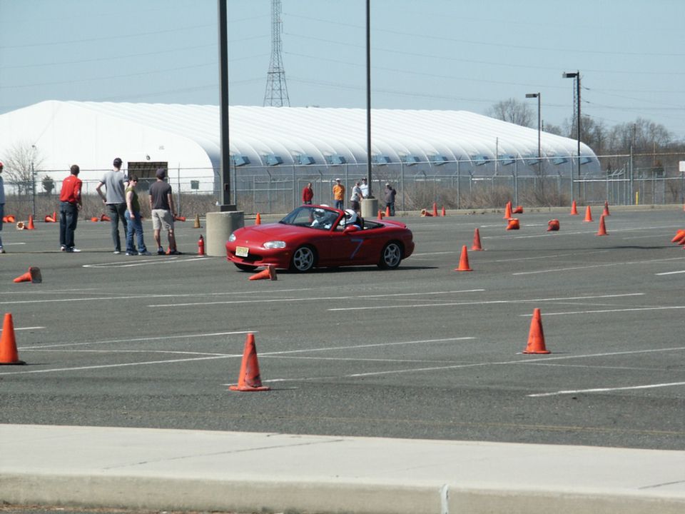 Red Mazda Miata convertible with racing number 7 on door, driving through an autocross course marked by orange cones