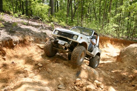 1990 Suzuki Samurai - A white off-road vehicle navigates a challenging dirt trail through a wooded area.