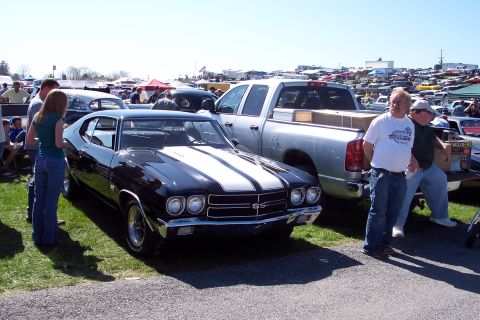 2001 Ford Crown Victoria - A classic black muscle car with white racing stripes is displayed at a car show, with a modern pickup truck and other vehicles in the backg…