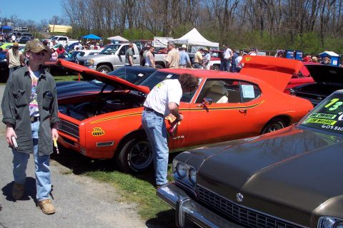 2001 Ford Crown Victoria - A man polishes an orange muscle car at an outdoor car show with other vehicles and people around.