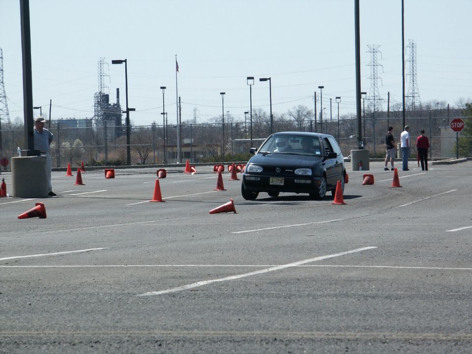 Black hatchback car on a paved lot with orange cones, driver visible
