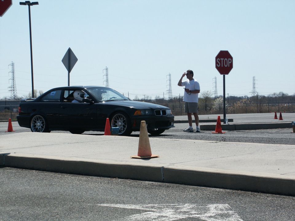 Black BMW E36 M3 with driver in helmet at an autocross event, cones and stop sign visible