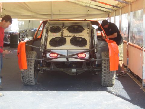 1990 Ford Bronco - Two crew members work on the rear of an orange-fendered off-road race truck fitted with three cooling fans under a tent.