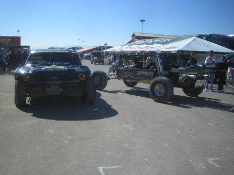 1990 Ford Bronco - black Ford trophy truck with Skyjacker and Toyo Tires graphics sits beside an open tube-frame buggy in a sunny paddock.