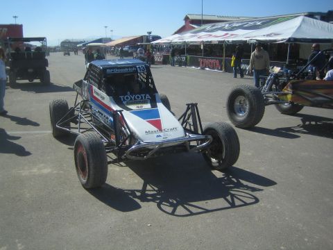 1990 Ford Bronco - silver-and-blue No. 13 tube-chassis off-road racer with Toyota and BFGoodrich graphics sits in a paddock under blue sky.