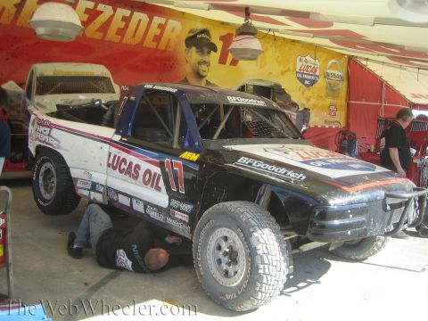 1990 Ford Bronco - black-and-white No. 17 Lucas Oil off-road race truck sits in a pit while a mechanic lies working beneath it.