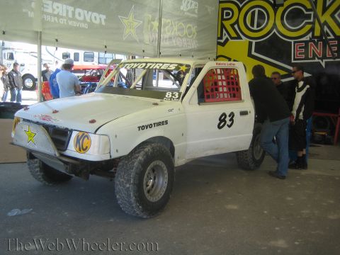 1990 Ford Bronco - white No. 83 off-road race truck with Toyo Tires, KC and Rockstar graphics sits in a pit as crew gather alongside.