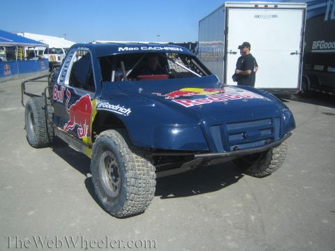 1990 Ford Bronco - dark blue Red Bull-liveried off-road race truck sits in a paddock with a man standing by an enclosed trailer behind it.