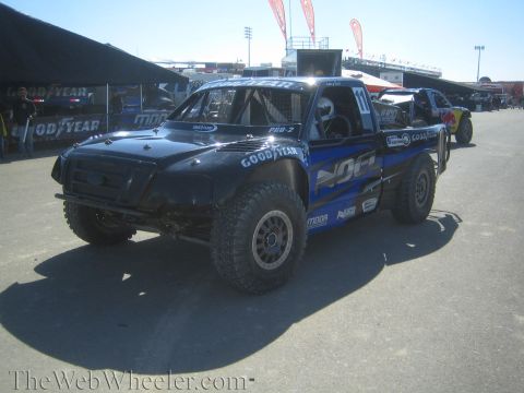 1990 Ford Bronco - black-and-blue off-road race truck with Goodyear and MOA graphics sits in a paddock with grandstands in the background.