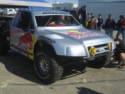 1990 Ford Bronco - silver Red Bull off-road race truck with a Ford grille badge, KC lights and BFGoodrich tires draws onlookers in a paddock.