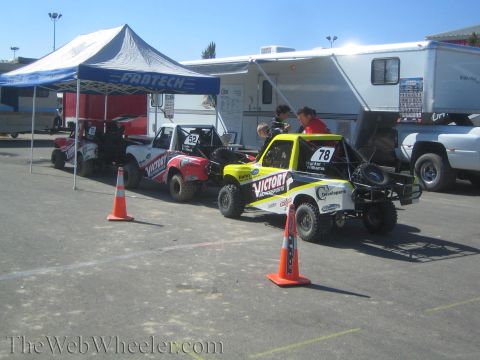 1990 Ford Bronco - yellow No. 78 off-road buggy and other race trucks line up beside a Fabtech canopy and RV with safety cones in the lot.