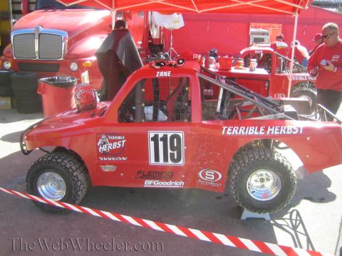 1990 Ford Bronco - red number 119 Terrible Herbst off-road race truck shown in side profile with exposed tube chassis and BFGoodrich graphics in a pit area.