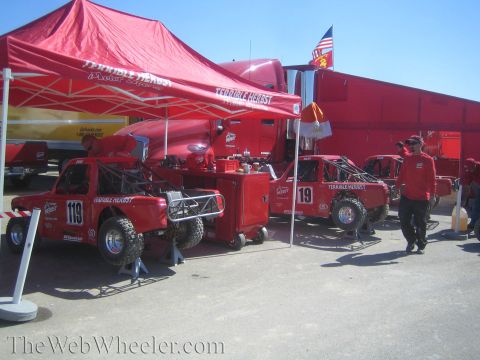 1990 Ford Bronco - Two red Terrible Herbst off-road race trucks numbered 119 and 19 sit in a pit area beside a large red hauler with an American flag overhead.