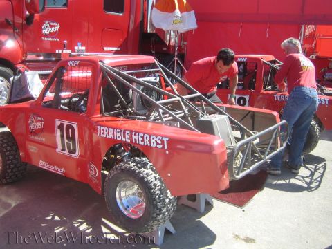 1990 Ford Bronco - Two crew members in red shirts work on the exposed rear tube chassis of a red number 19 Terrible Herbst off-road race truck in the pits.