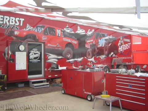 1990 Ford Bronco - interior of a Terrible Herbst race hauler showing a graphic-wrapped wall of trucks and red rolling tool cabinets under a tent.