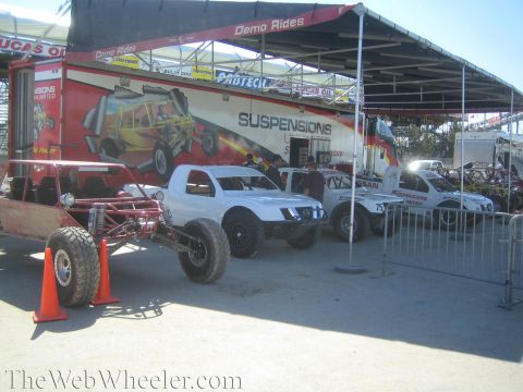 1990 Ford Bronco - Several white and silver off-road race trucks and a tube-chassis buggy parked under a canopy beside a Suspensions display trailer.