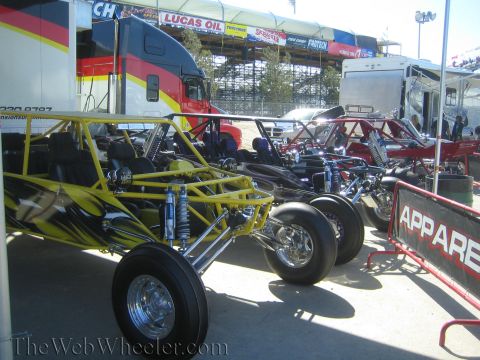 1990 Ford Bronco - yellow tube-chassis off-road buggy with large rear tires parked in a pit area among other vehicles and race haulers.