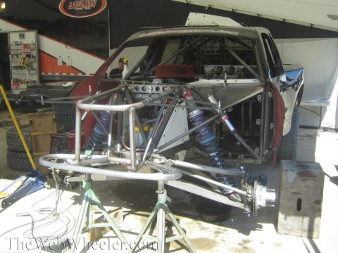 1990 Ford Bronco - off-road race truck with its front body panels removed, exposing the tube chassis and blue coilover shocks, sitting on jack stands in a sho…
