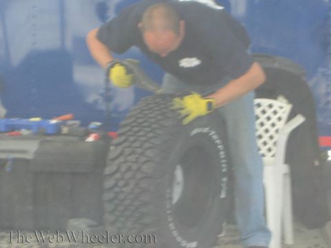 1990 Ford Bronco - man wearing yellow work gloves leans over to mount a Mickey Thompson off-road tire in a blue-walled work area.
