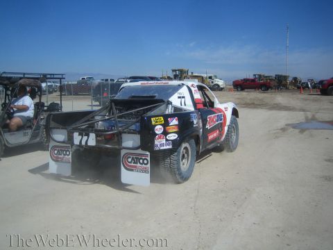 1990 Ford Bronco - black and white number 77 off-road race truck with CATCO mud flaps parked on a dirt lot with heavy equipment in the background.